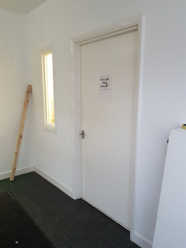 Hallway with the white painted door on which the drawing is cellotaped at eye level, light comes through a tall slot window to the left of the door, a length of wood leans against the corner of the wall left of the window