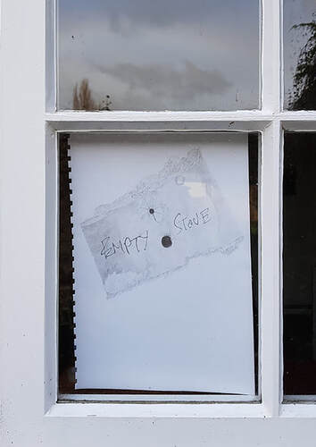 Close up of a small window pane within a white painted frame, through the window is an A4 sheet of white paper torn from a notebook affixed from behind almost filling the window, on the paper an accurate drawing of a torn piece of card with two holes above one another in the centre and roughly handwritten the word 