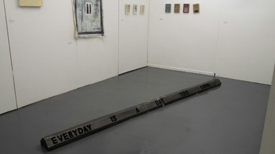 Two black car stops end to end with the enscribed words "Everyday is a day too long" on the grey floor of a gallery with white walls with small paintings hung