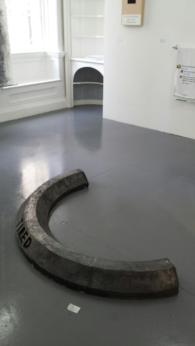 Black crescent shaped car stop with enscribed word "Tired" on grey floor of a gallery with white walls