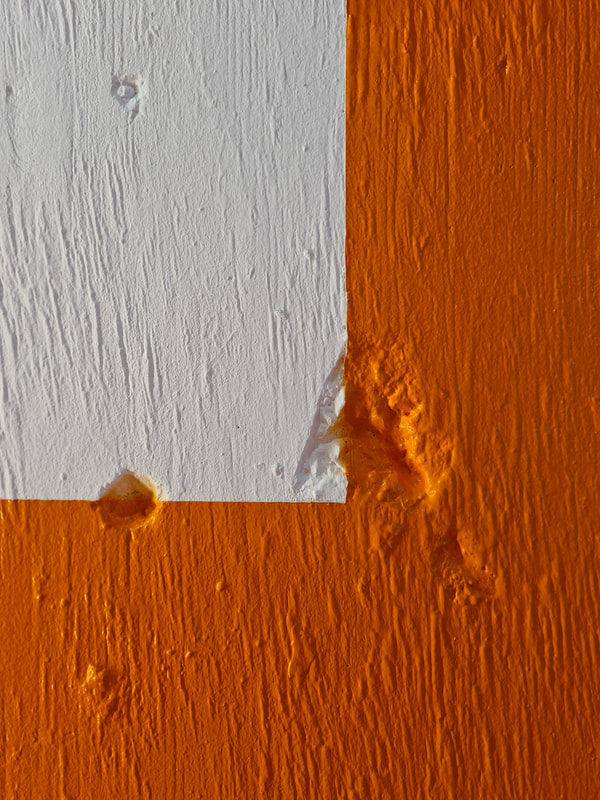 Bottom right corner of white portrait orientated rectangle on orange painted concrete wall. The wall is textured with lines and pits. The orange boarder intersects two deep pits on the bottom right corner of the rectangle. 