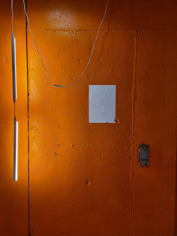 White portrait orientated rectangle on orange painted concrete wall. The wall is textured with lines and pits. To the left are two white strip lights hanging from a cable illuminating the orange wall. To the right is a small sator square woven in thread into a small piece of tree bark mounted on the wall.