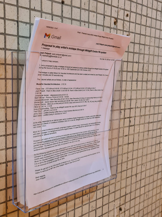 Printouts of email with Gmail logo in top left corner of sheet in an upright stack sitting in a clear plastic document holder mounted to a white tiled wall.