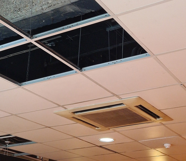 White tiled ceiling with square air conditioner unit. The air conditioner has slotted air vents and some of the ceiling tiles are missing, showing concrete beams and metal fixings beyond.