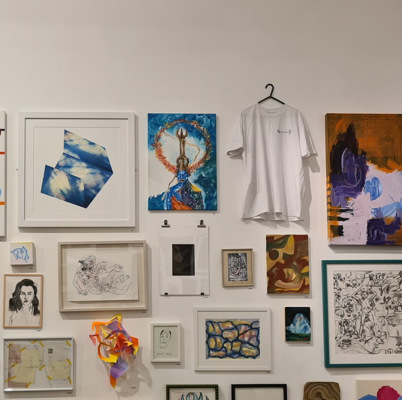 White t-shirt with black double-headed arrow embroidered on the left breast, hung on a clothes hanger high on a white art gallery wall amongst various paintings and drawings.