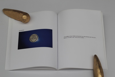 A book held open on a white table with golden skatestops as paper weights, left page reads "Dearrrr!" in all capitals above a picture of a love heart sweet which reads "Yes Dear" on a dark blue surface with a camera flash reflection, right page reads "I am willing to buy chicken and prevent you from disease and I buy you flowers. It's safe to say I love you too potato"