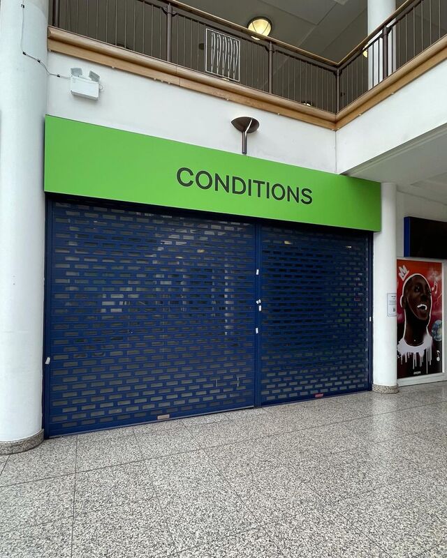 Blue shop window shutters with small art stickers stuck along the frame four up the centre and one to the left, above the shutters a green banner with centered black capitalised text reading "conditions" 
