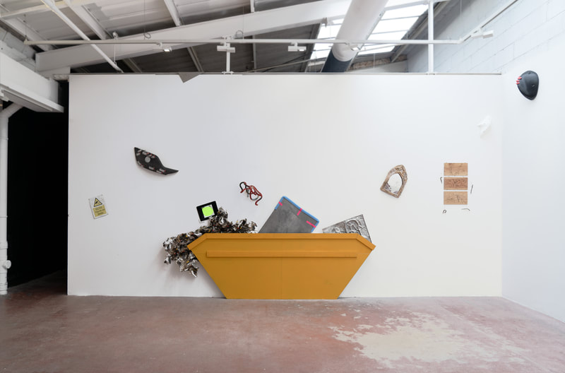 White art gallery wall with a yellow painted wooden cut out prop resembling a rubbish skip centered on the floor. Multiple art works have been mounted behind and above the prop as if art is spilling out of the skip. Art works are also mounted scattershot across the wall.