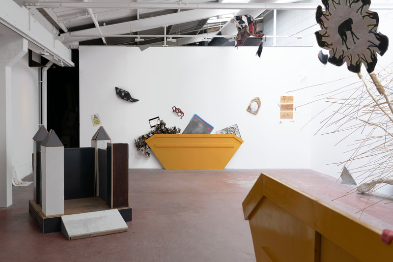 White art gallery wall with a yellow painted wooden cut out prop resembling a rubbish skip centered on the floor. Multiple art works have been mounted behind and above the prop as if art is spilling out of the skip. Art works are also mounted scattershot across the wall. In the foreground a sculpture of a castle made from cut wooden hoarding. 