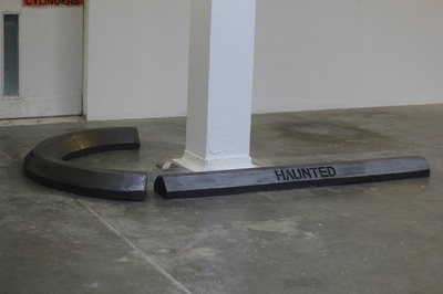 Black crescent shaped car stop and black car stop with enscribed word "Haunted" end to end to make a hook shape on grey floor around white square column