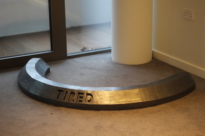 Black crescent shaped car stop with enscribed word "Tired" on grey carpet in front of round white column 