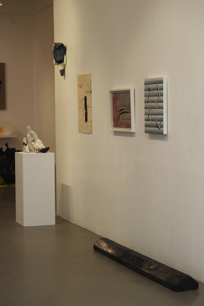 Black car stop with enscribed word "Haunted" on grey floor next to a sculpture on a white plinth parrallel to a white wall with three paintings hung on it