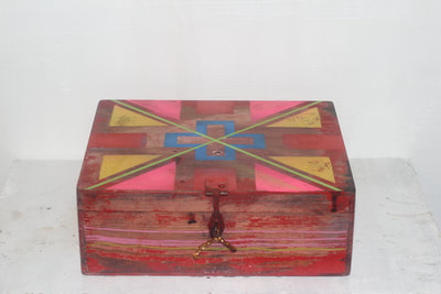 Red wooden box resting on a white shelf, closed with a blue medical cross, yellow and pink triangles, red squares, and a green X painted on the lid