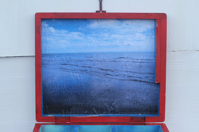 Picture of calm blue sea under clear sky with puffy couds pasted under the open lid of the red box