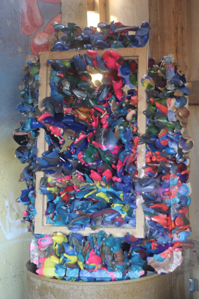 An abstract sculpture with splotches of blue and purple and pink and green in and around a wooden picture frame hanging in the air
