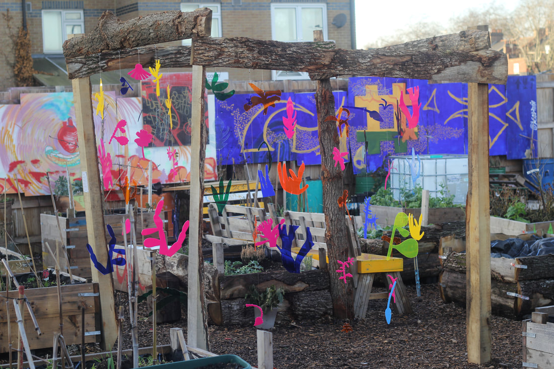 A large frame with lengths cut from tree trunks with bark standing in an urban community garden, many small plastic cut outs of different plants coloured yellow, pink, blue, green, and orange hang from wires on the frame
