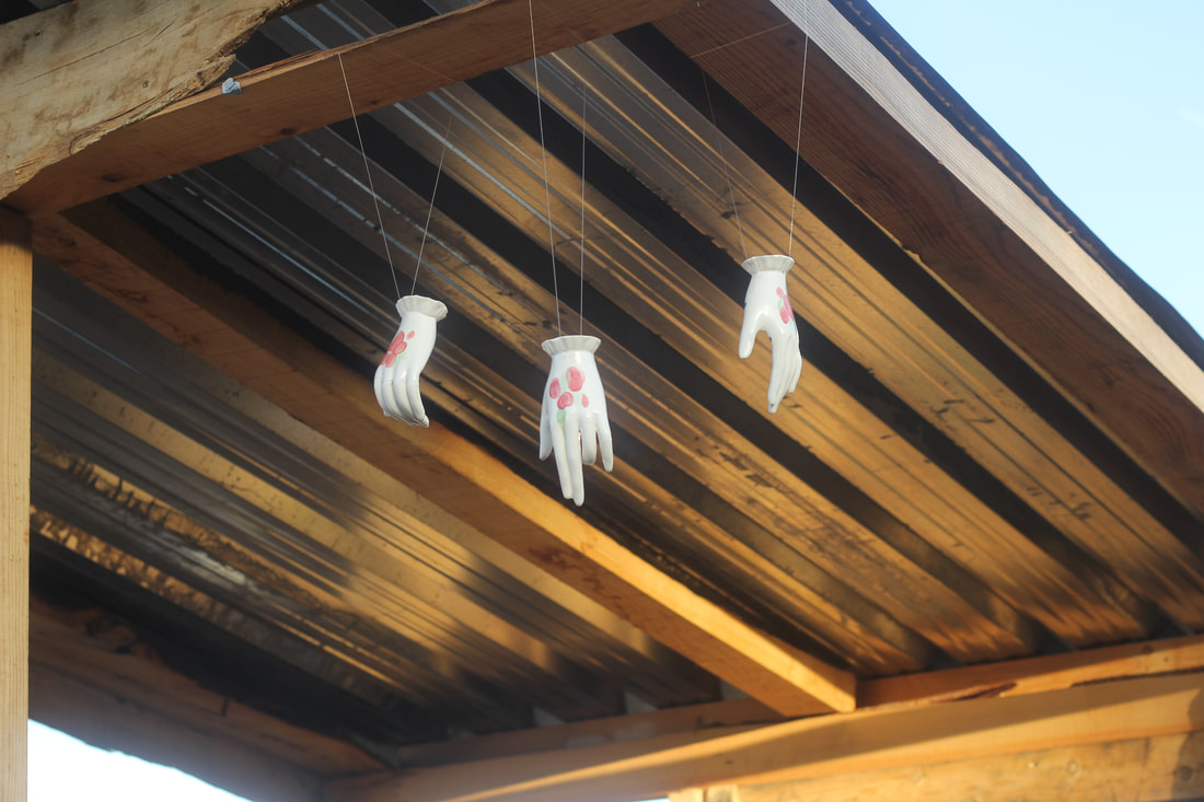 Three white ceramic hands each with glazed illustrations of red berries on their backs hang by the wrists from wires attached to a wooden structure with a corrugated metal roof