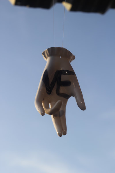 A white ceramic hand with a slightly bent ring finger and "VE" glazed in black on its palm hanging by its wrist on a wire
