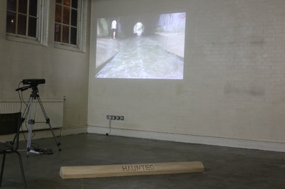 Plaster cast car stop with enscribed word "Haunted" on grey floor in white walled room with a film projected onto the nearest wall